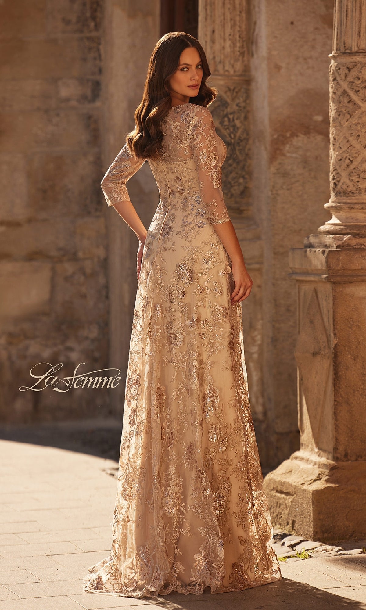 Wearing the La Femme 30803, a long floral-lace formal dress with three-quarter sleeves, a woman stands outdoors by stone columns, facing away but glancing over her shoulder—perfect for the mother of the bride.